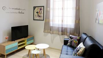 A living room with a TV on a wooden cabinet, a black sofa with patterned cushions, and a small white table.