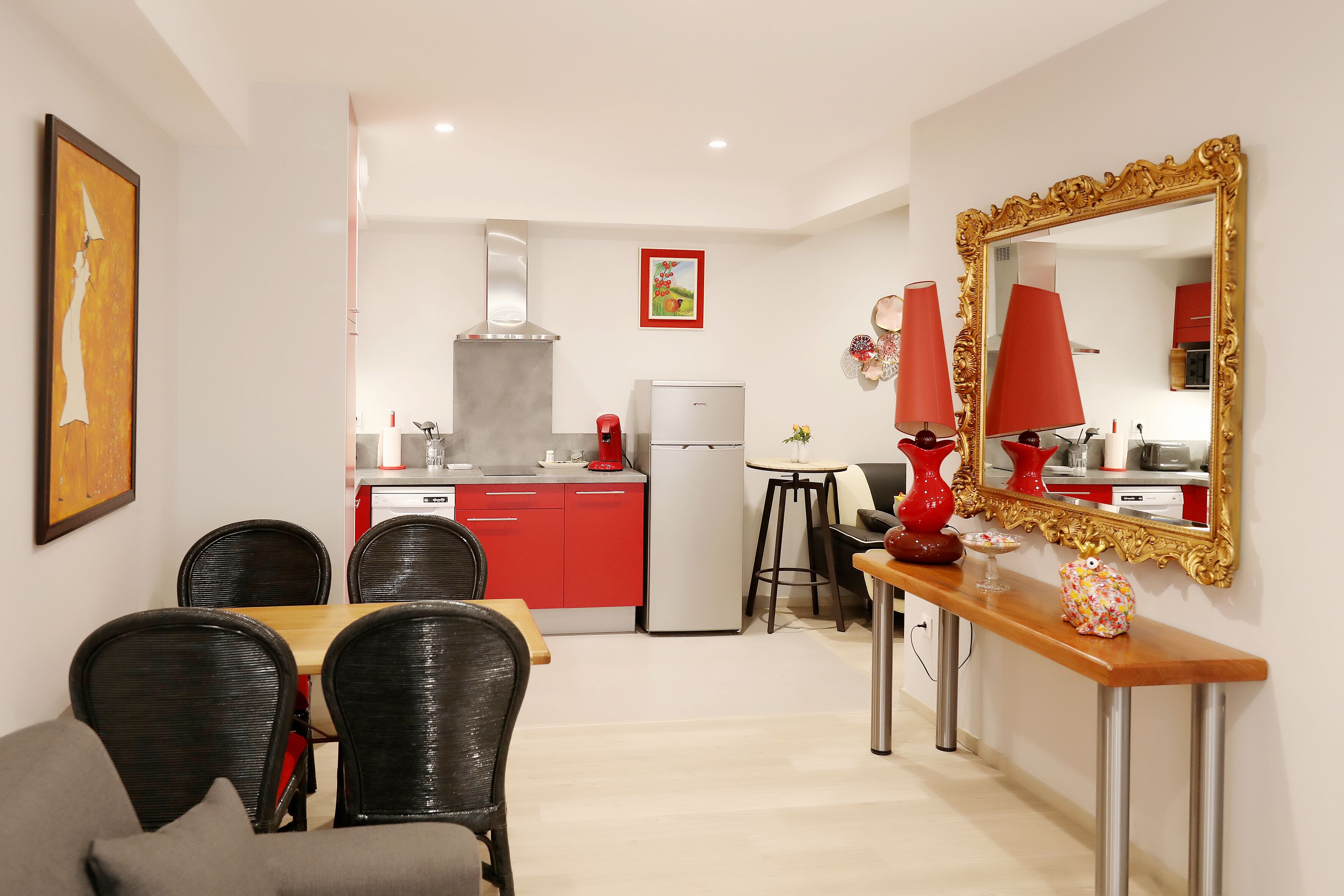 A modern kitchen with red cabinets, a white refrigerator, and a stainless steel range hood. There is a dining area with a wooden table and black chairs. A large mirror with a gold frame is on the wall.
