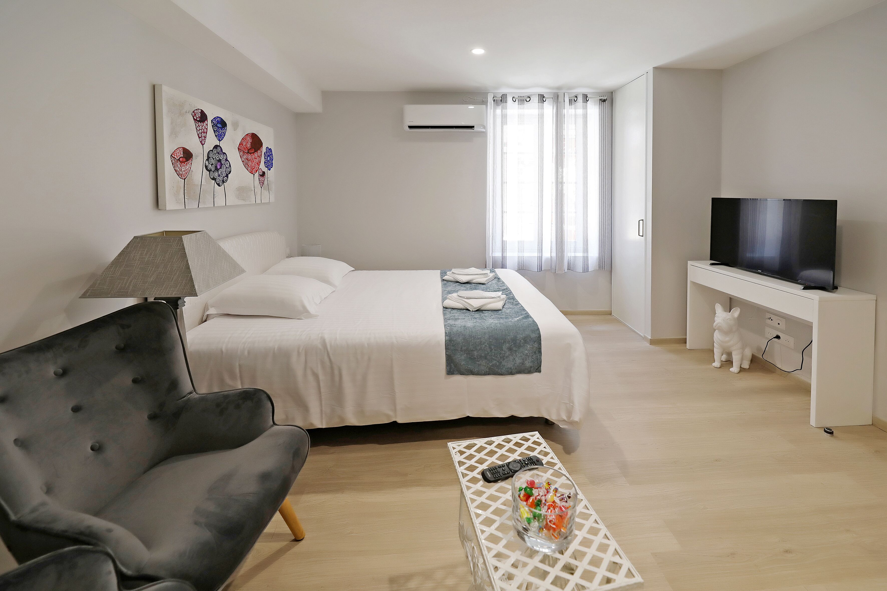 A modern hotel room with a bed, a TV, a chair, and a small table with a remote control and a glass of candies.
