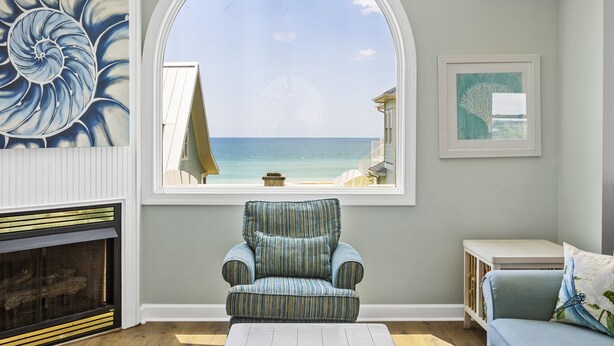 A room with a large arched window overlooking a beach, a striped armchair, a sofa, and a fireplace. - Sea La Vie | Oceanfront + Community Pool! (North Topsail Beach)