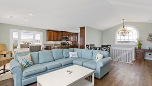 A spacious living room with a light blue sectional sofa, a white coffee table, and a dining area with a white railing. - Sea La Vie | Oceanfront + Community Pool! (North Topsail Beach)