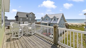 A wooden deck with white chairs and a table, overlooking a beach with houses in the background. - Sea La Vie | Oceanfront + Community Pool! (North Topsail Beach)