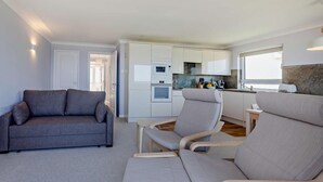 Un espace de vie compact comprenant un canapé gris, un coin repas avec une table et des chaises, un coin cuisine et une fenêtre laissant entrer la lumière naturelle.