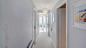 Un couloir étroit aux murs blancs, avec un radiateur et un luminaire au plafond. Des portes donnent accès à d’autres pièces, et une œuvre d’art encadrée est accrochée au mur de droite.