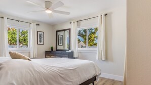 Un dormitorio con cama, ventilador de techo, cómoda y dos ventanas con cortinas.