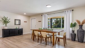 Un comedor con mesa y sillas de madera, un armario negro y un ventanal con cortinas blancas.