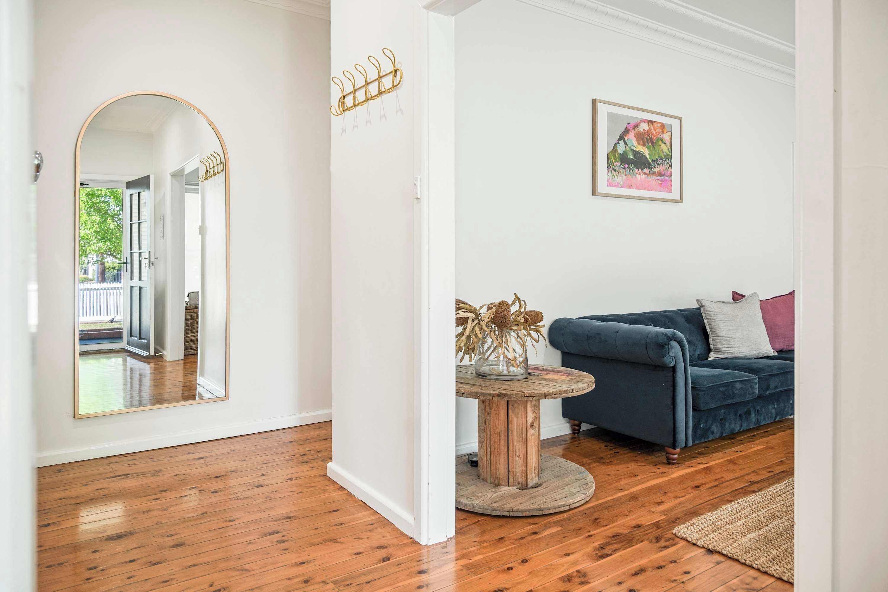 Una habitación con piso de madera, una mesa redonda de madera con un florero, un sofá azul y un cuadro enmarcado en la pared.