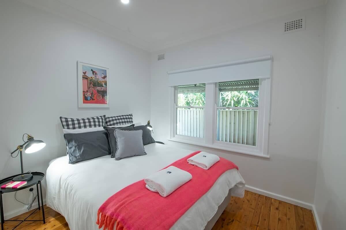 Un dormitorio con cama, almohadas, lámpara de noche, ventana con persianas y un cuadro en la pared.
