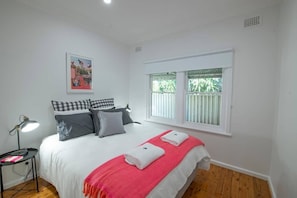 A bedroom with a bed, pillows, a bedside lamp, a window with blinds, and a painting on the wall.