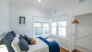 A bedroom with a bed, pillows, a teddy bear, a blue blanket, a white towel, a wall-mounted shelf, and a window. - Cobbora Cottage -  Sleek & Stylish, Near Hospital (Dubbo)
