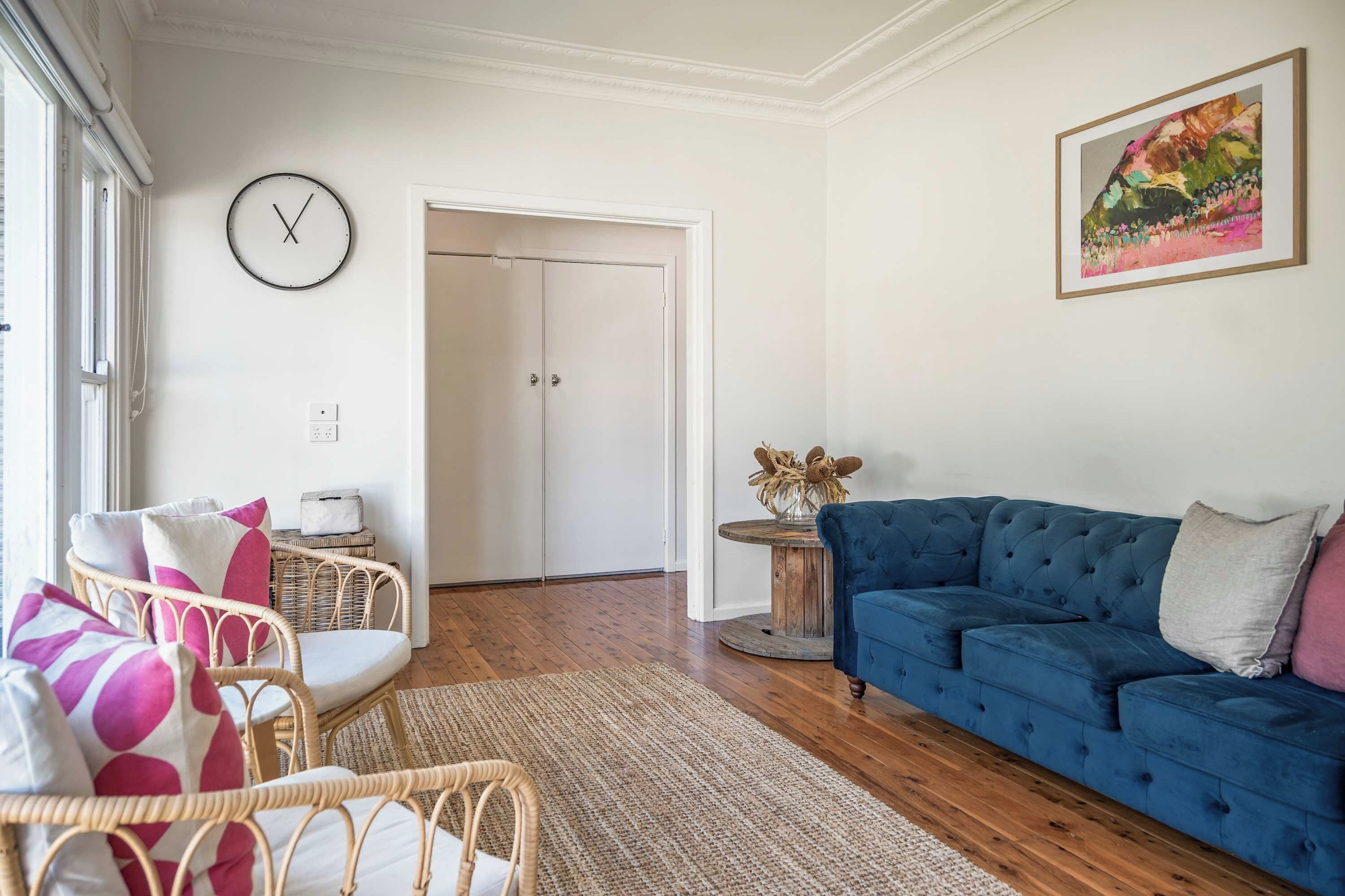 Olohuone, jossa on sininen sohva, rottinkituoleja, pyöreä sohvapöytä ja seinäkello.
