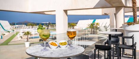 Een terras met een gedekte tafel voor twee personen, met een cocktail en een biertje, en een zwembad en palmbomen op de achtergrond.