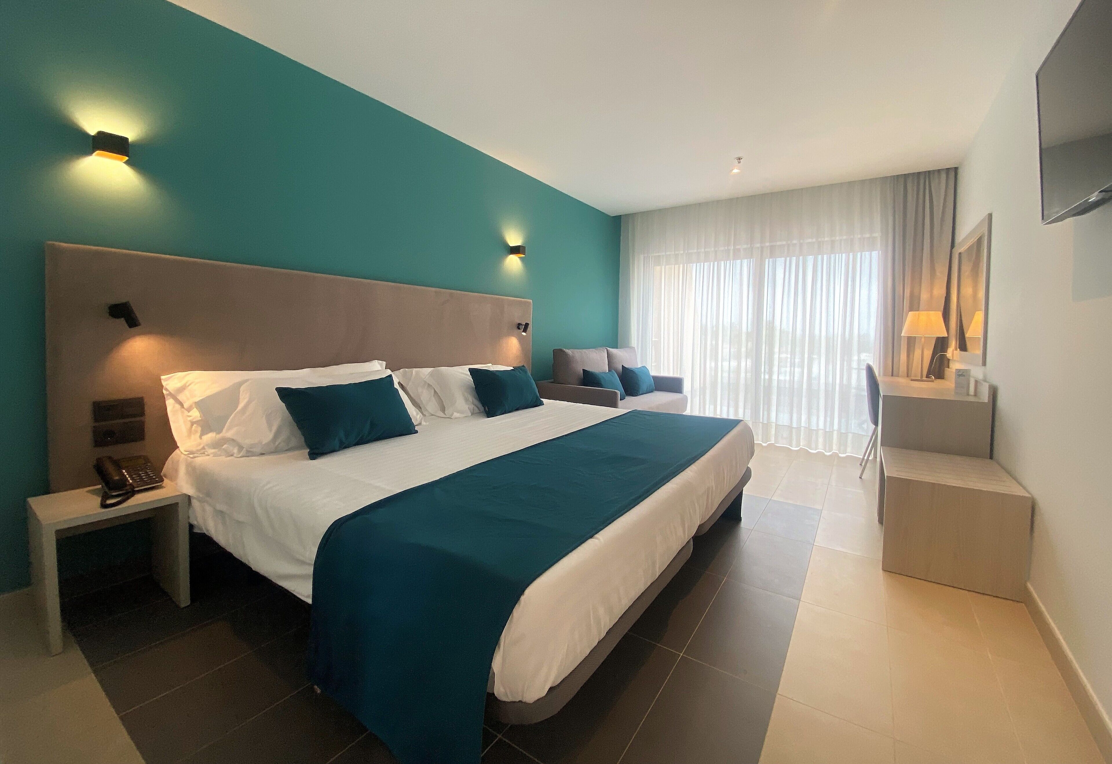 Una habitación de hotel moderna con una cama grande, dos mesitas de noche, un escritorio y un amplio ventanal con cortinas.