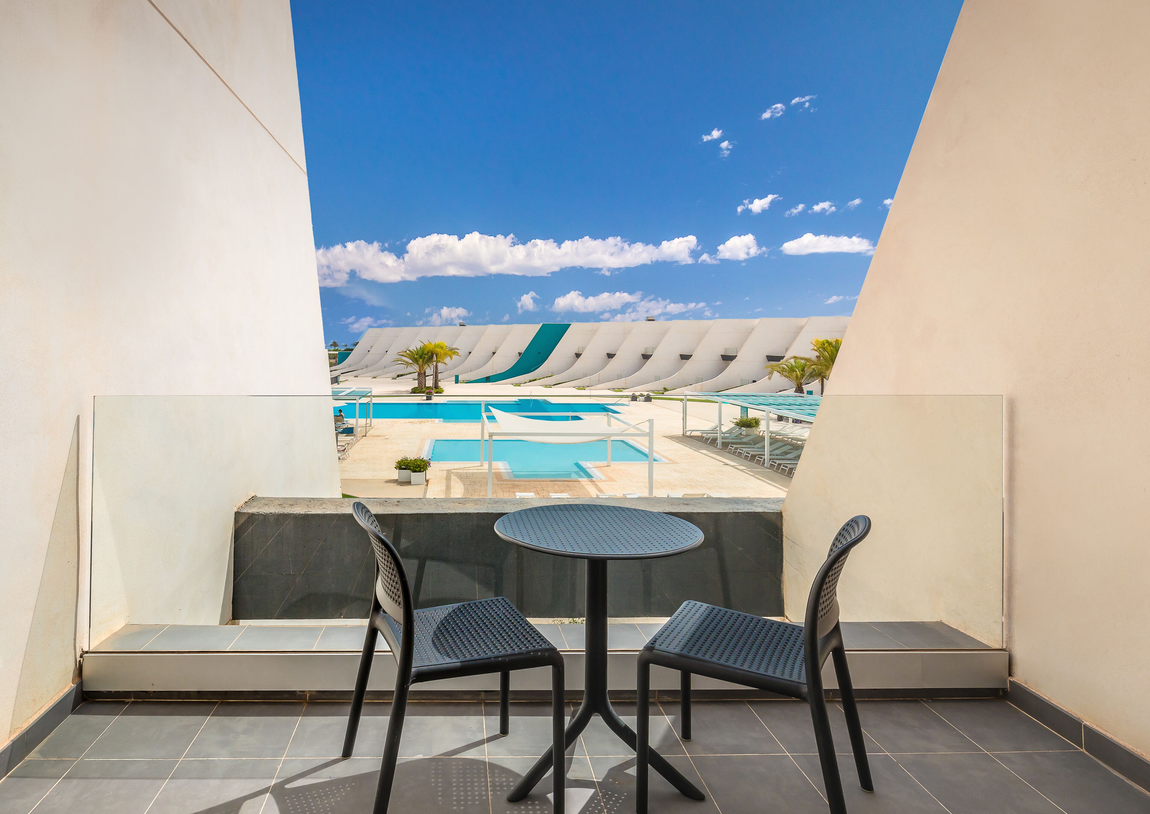 A balcony with two chairs and a small table, overlooking a pool area with multiple swimming pools and lounge chairs.