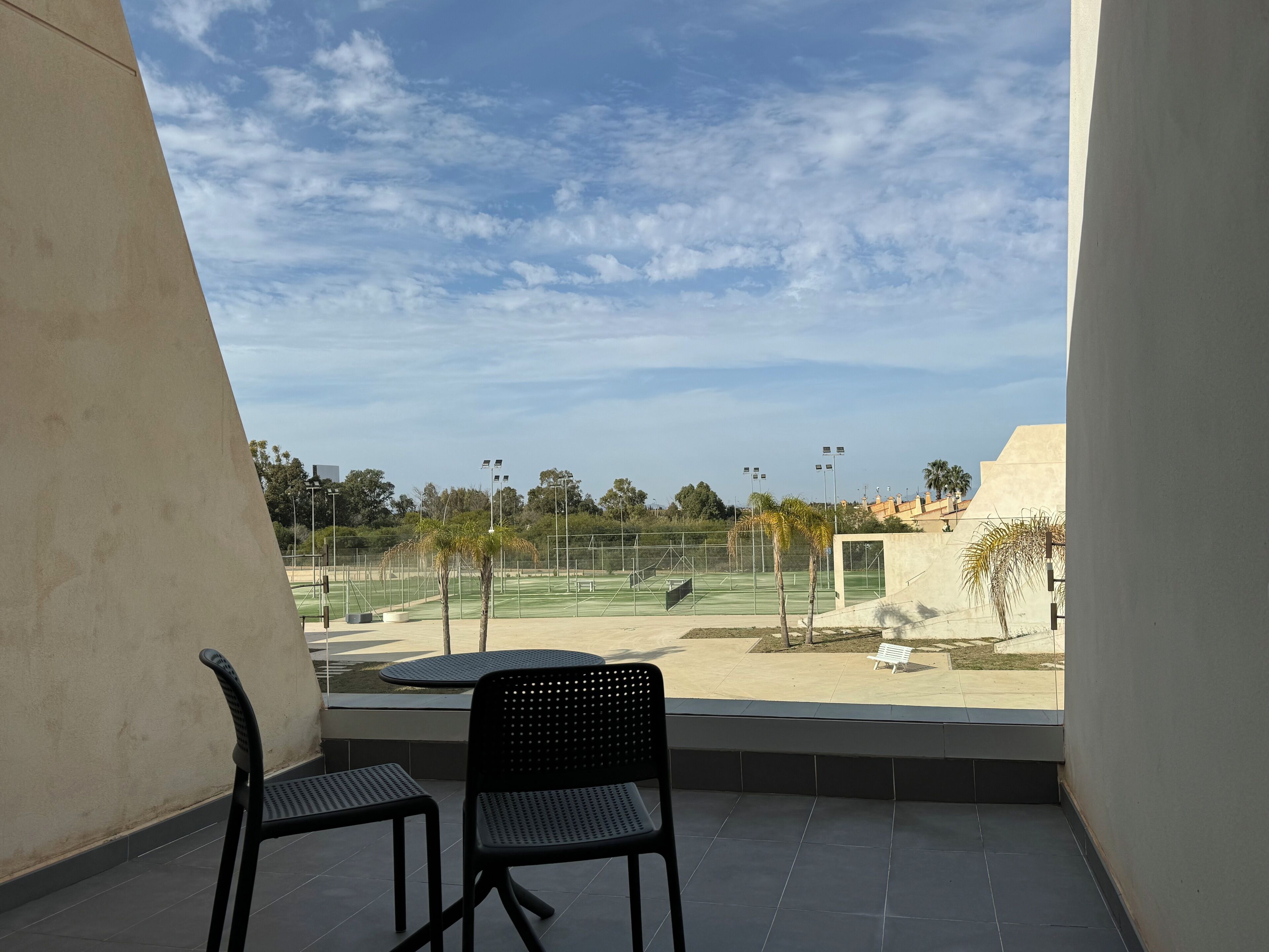 A balcony with two black chairs, a tennis court in the background, and a clear sky.