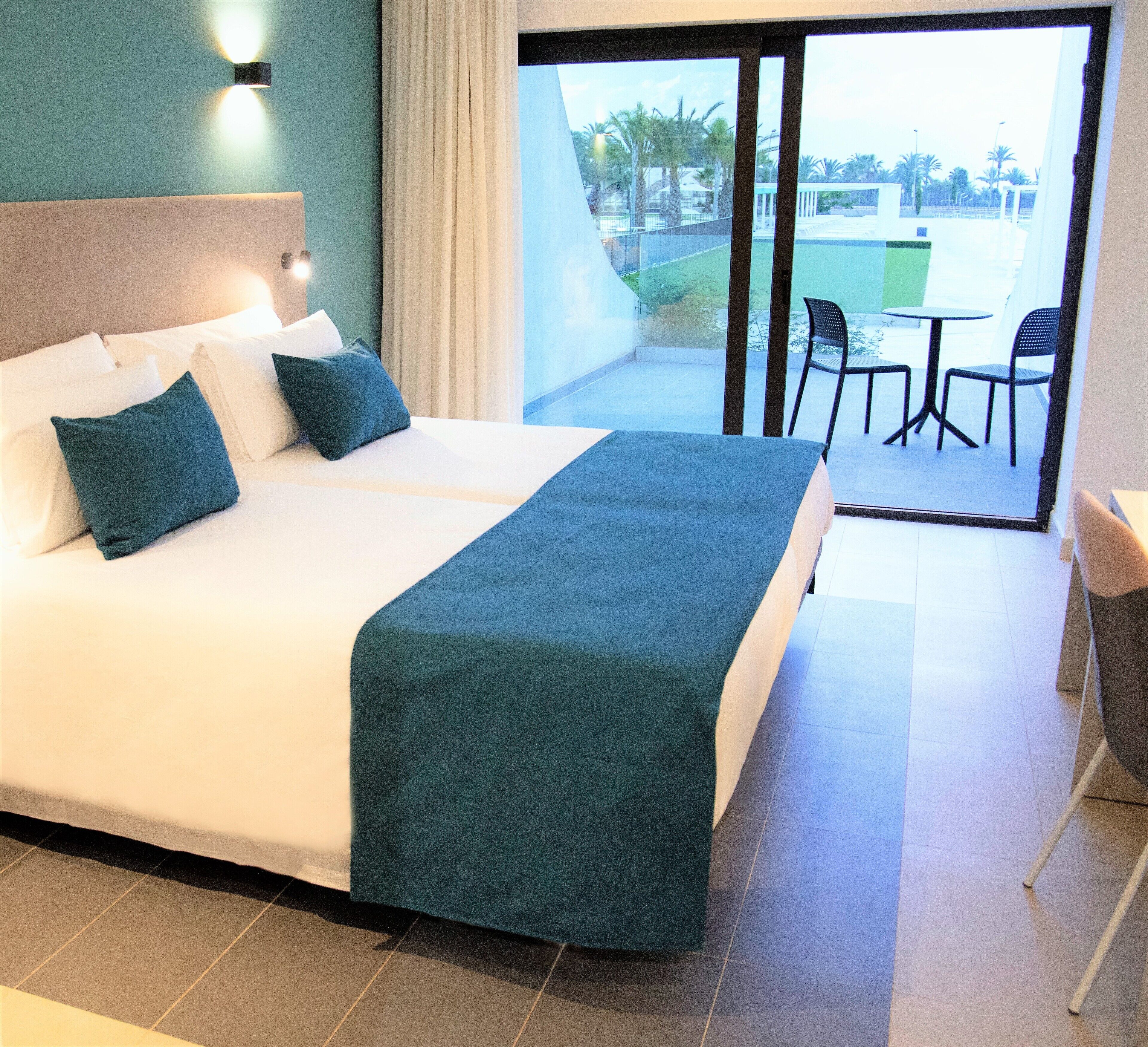A hotel room with a large bed, a view of the outdoor area through a sliding door, and a small table with chairs.