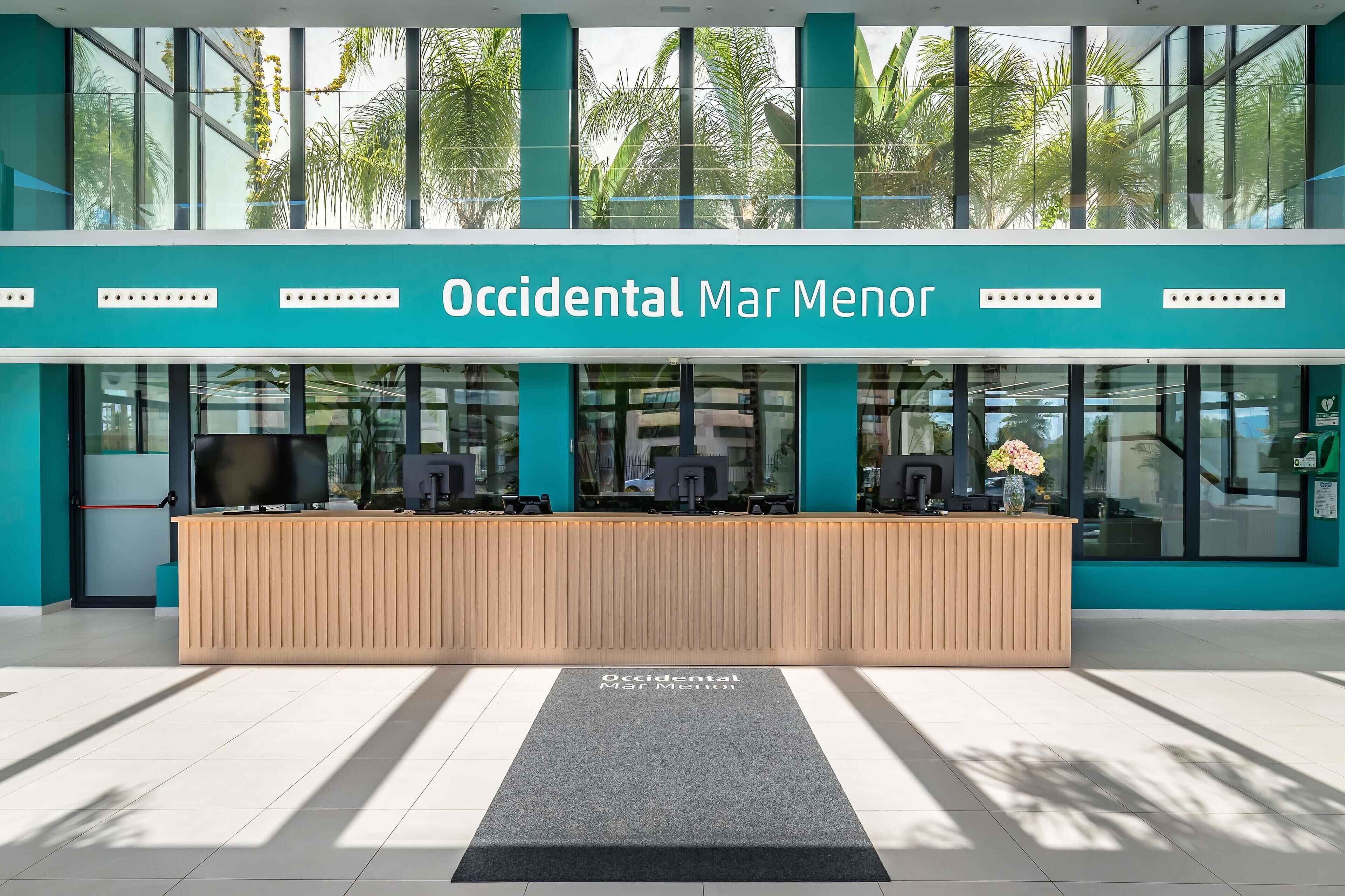Un comptoir d’accueil avec une enseigne indiquant « Occidental Mar Menor », situé dans un bâtiment moderne doté de grandes fenêtres et offrant une vue sur des palmiers.