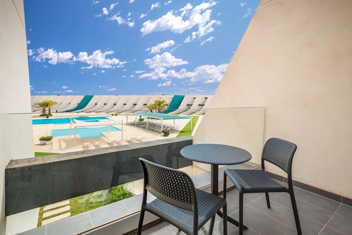 A balcony with two chairs and a table, overlooking a swimming pool and a building with a unique architectural design.