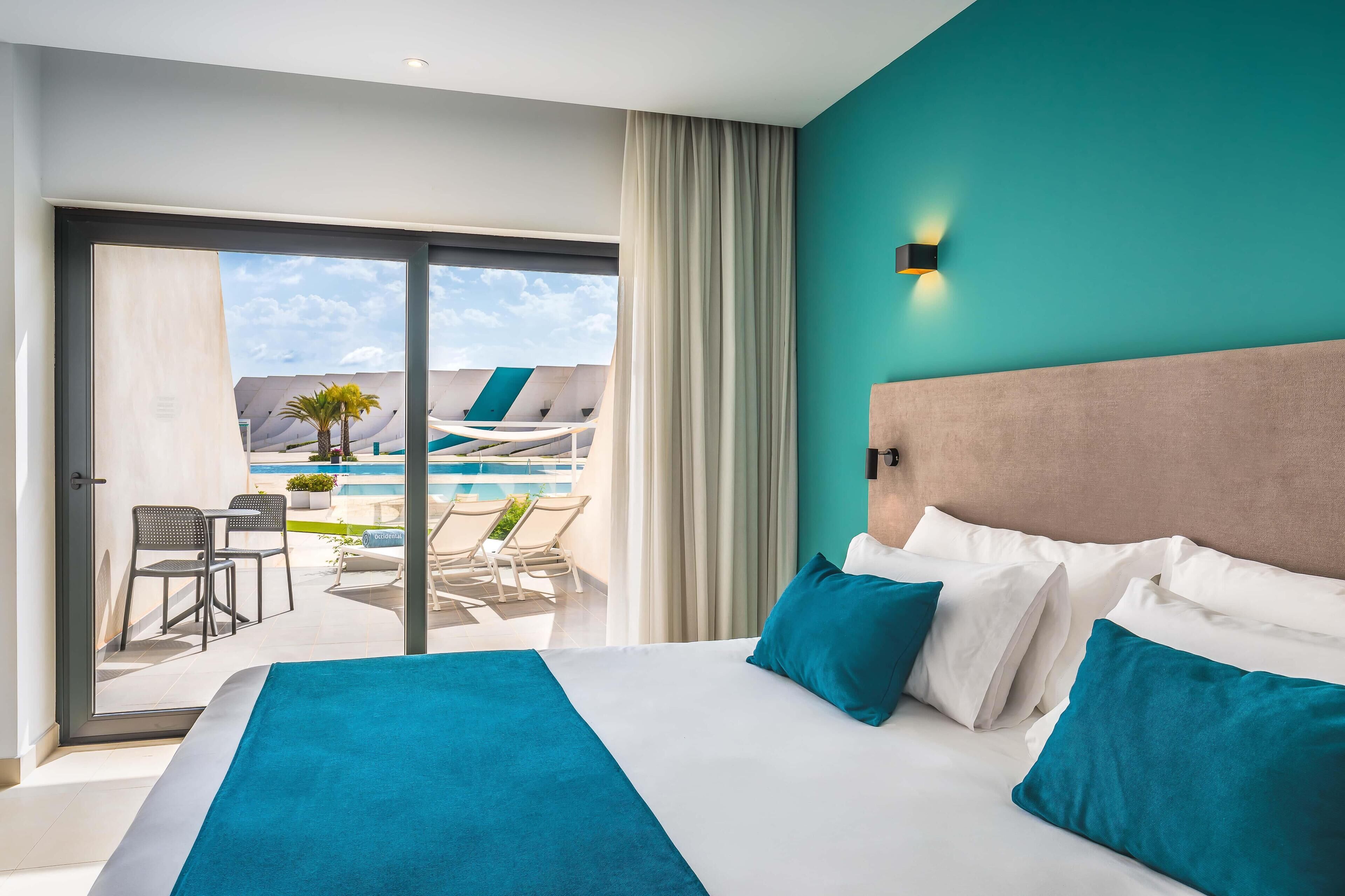 A hotel room with a large bed, a view of a pool and lounge area, and a teal accent wall.