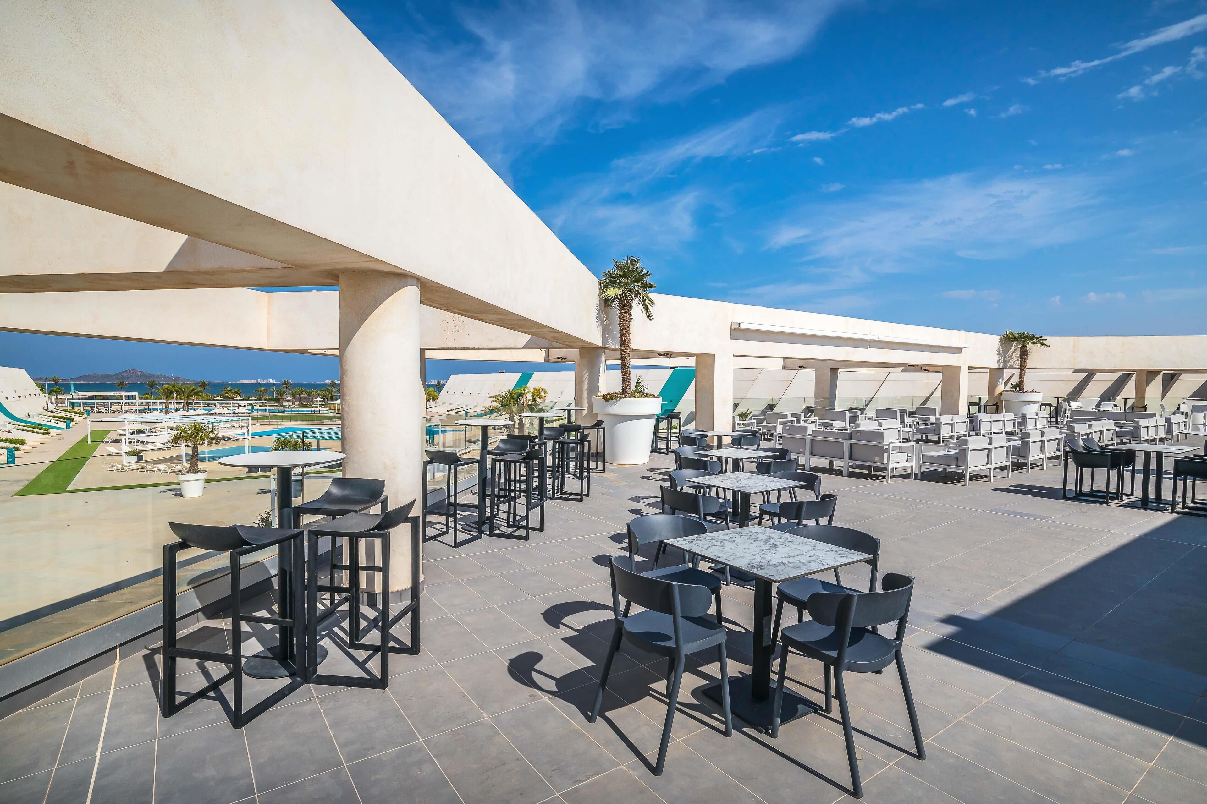 An outdoor terrace with tables and chairs, palm trees, and a view of the sea.