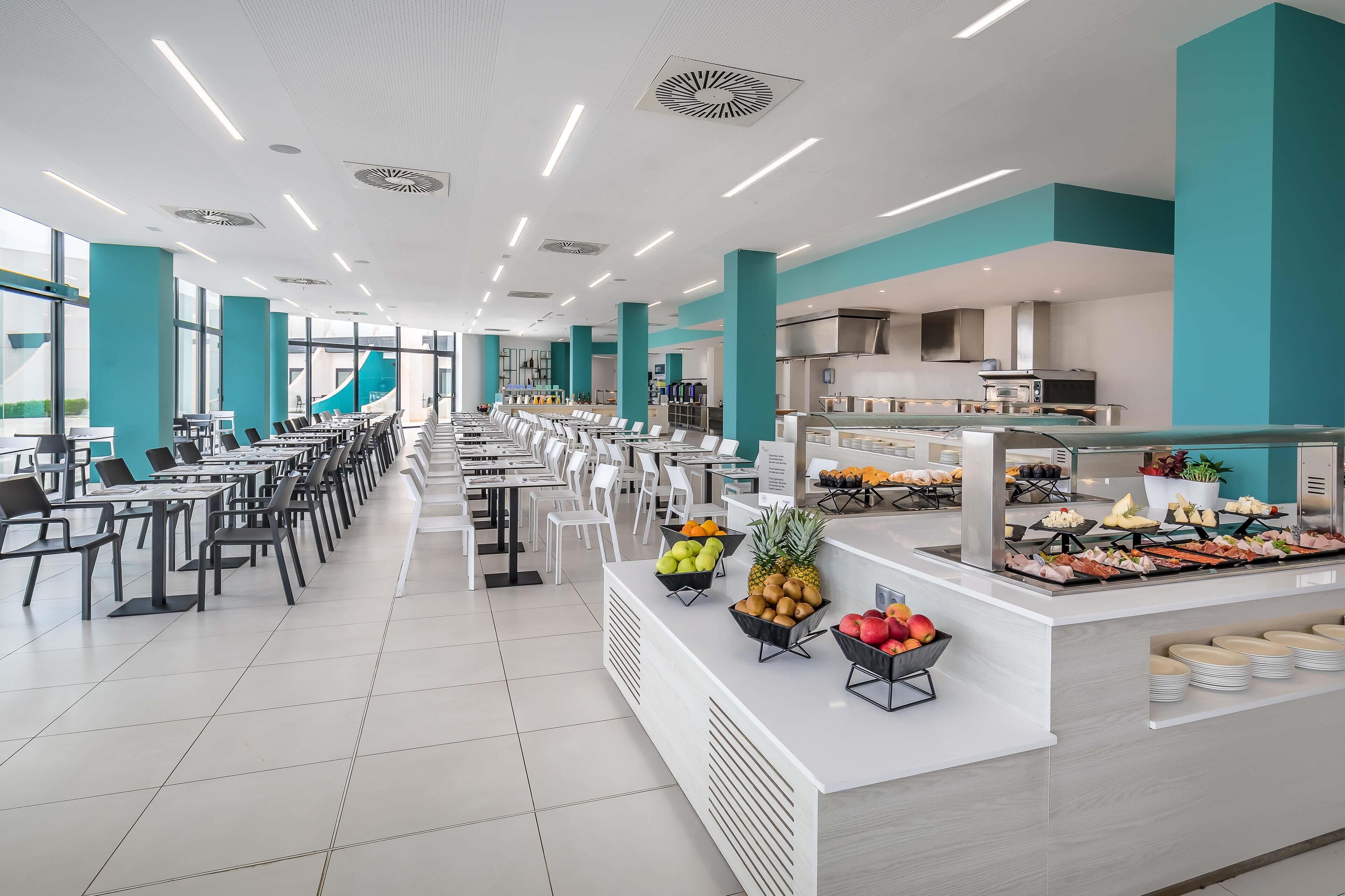 A modern cafeteria with long tables and chairs, a central food display with fruits and snacks, and a kitchen area in the background.