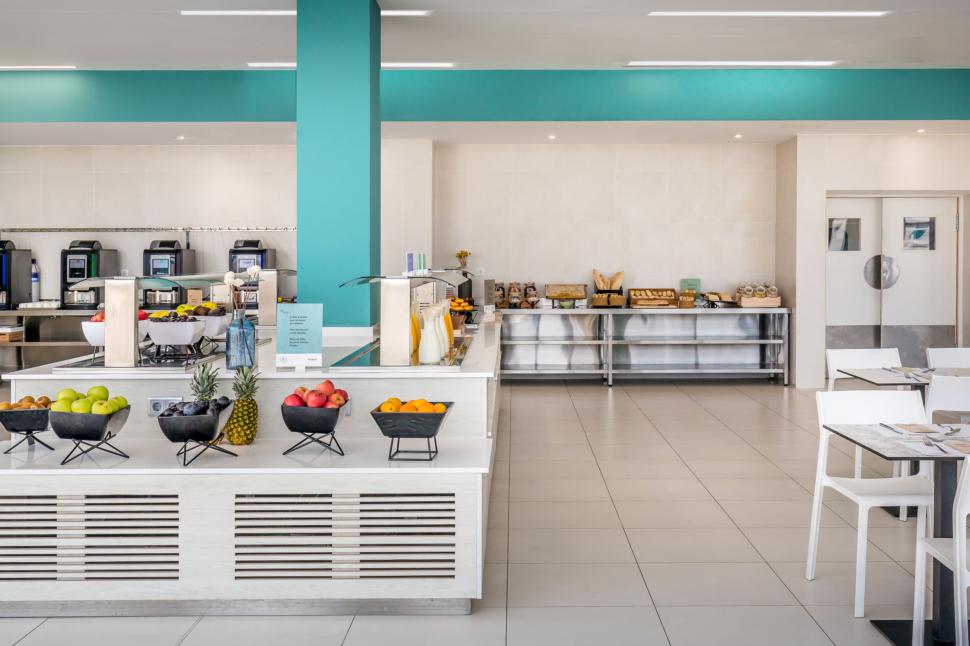 A modern cafe with a central counter displaying fresh fruits, a seating area with white chairs, and a teal-colored ceiling.