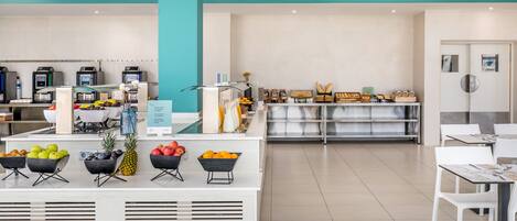 A modern cafe with a central counter displaying fresh fruits, a seating area with white chairs, and a teal-colored ceiling.