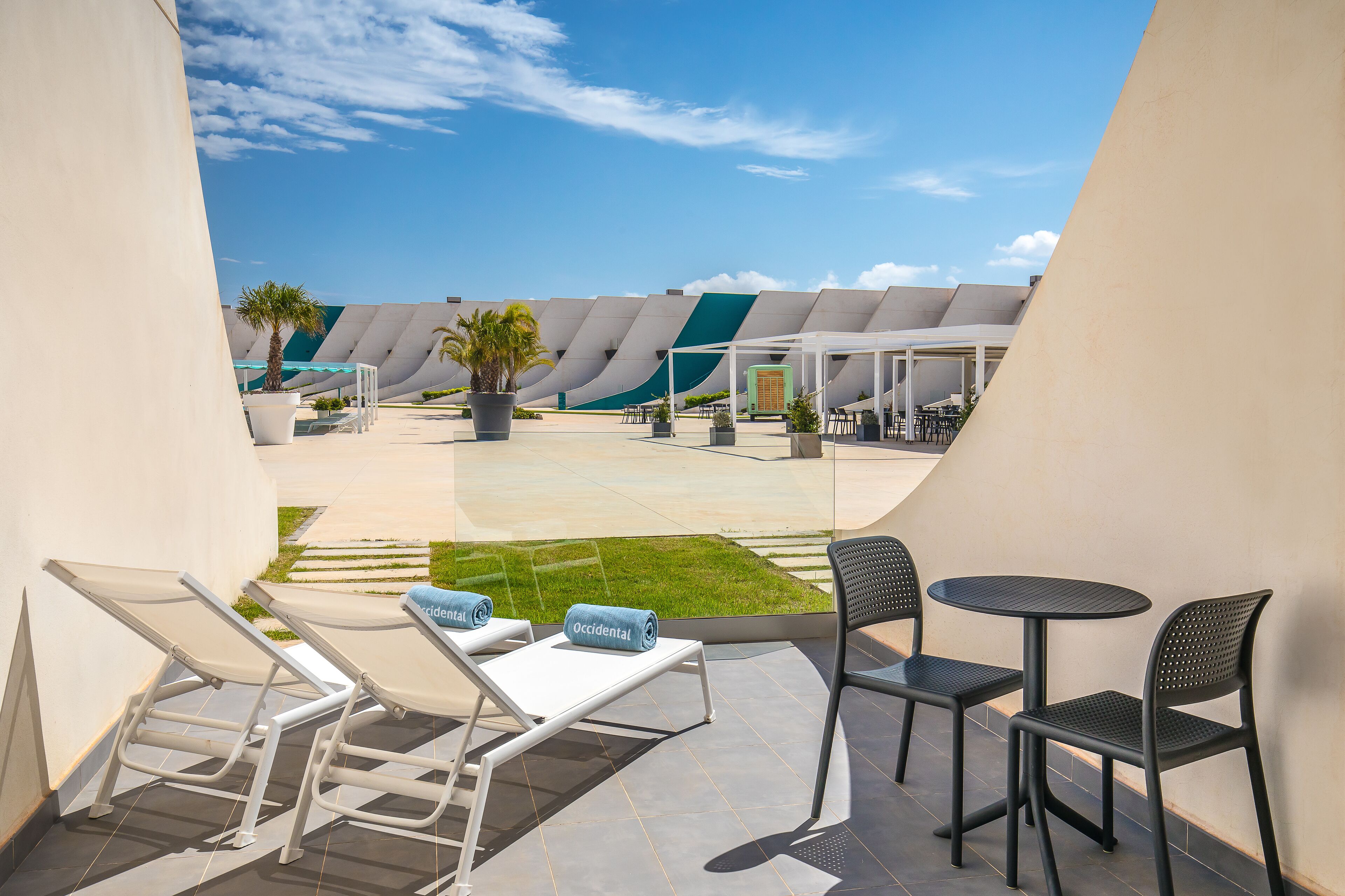 Un área de estar moderna al aire libre con sillones reclinables y mesas, un césped y un diseño arquitectónico singular al fondo.