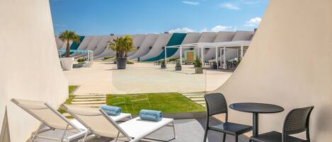 Un área de estar moderna al aire libre con sillones reclinables y mesas, un césped y un diseño arquitectónico singular al fondo.