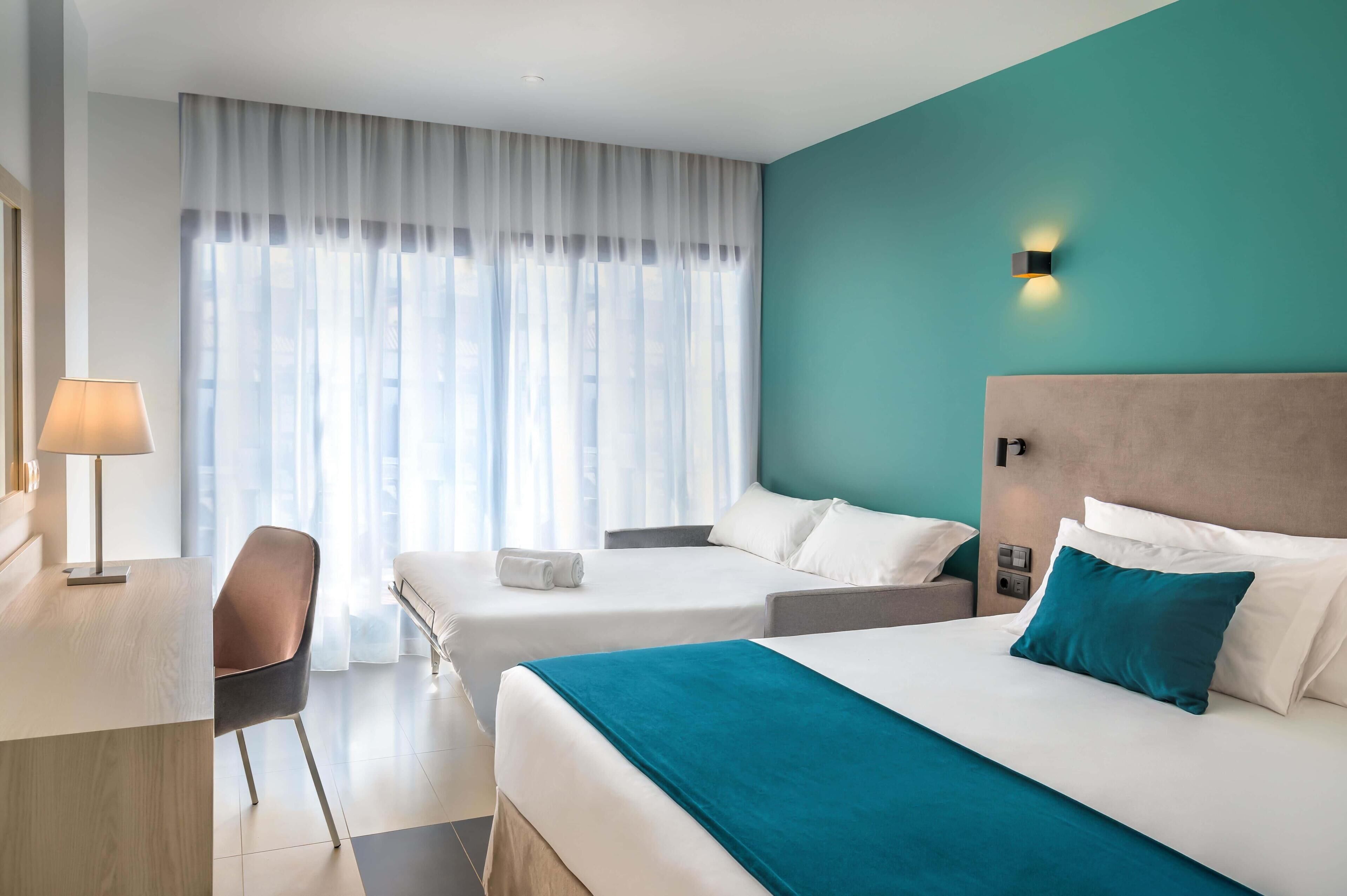 Habitación de hotel con dos camas, un escritorio, una silla y una ventana con cortinas traslúcidas.