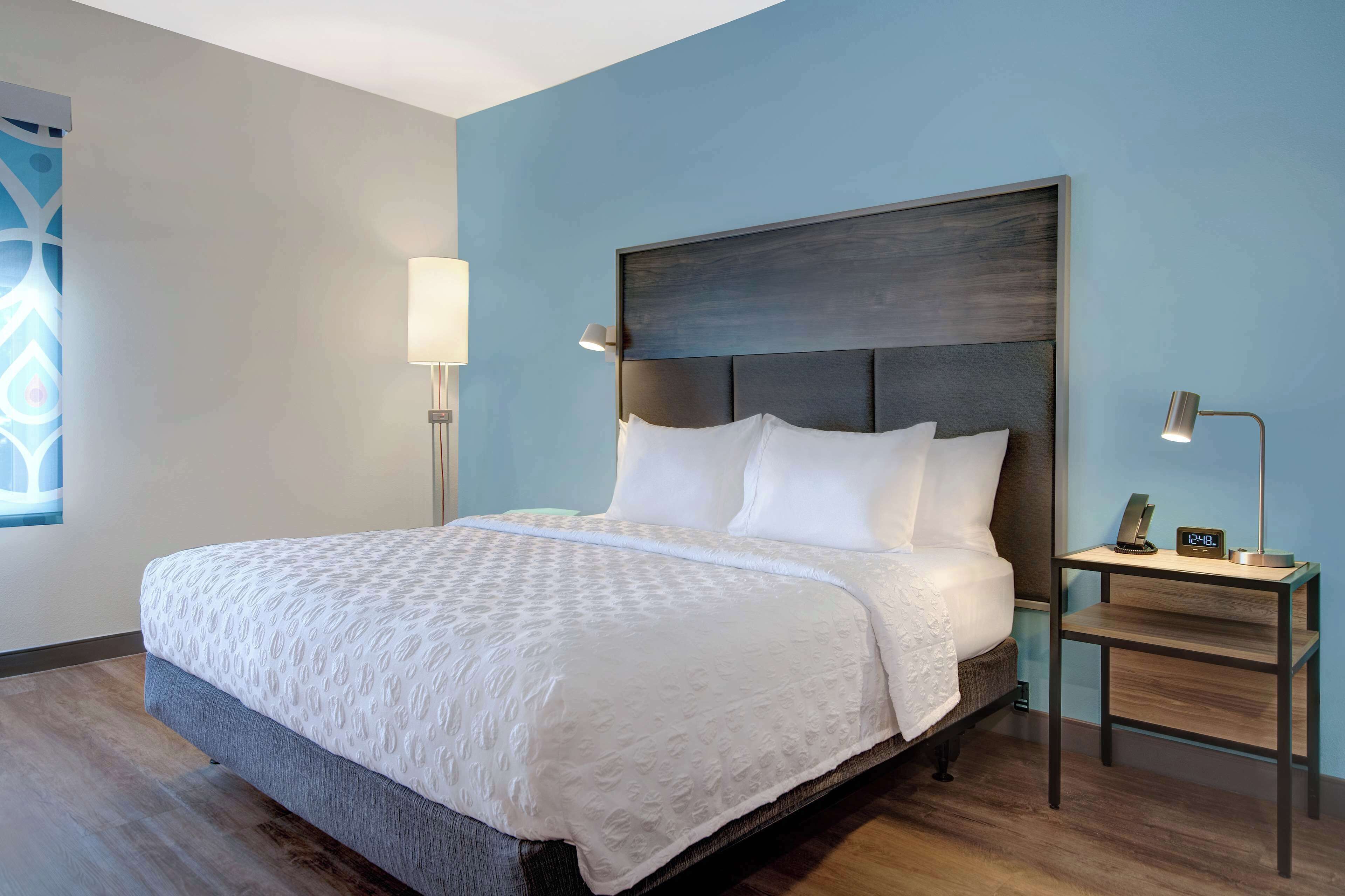 A neatly made bed with a white bedspread and pillows, a bedside table with a lamp, and a wall clock.