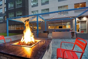 Un área de estar al aire libre con una chimenea, sillas rojas y un edificio moderno al fondo.