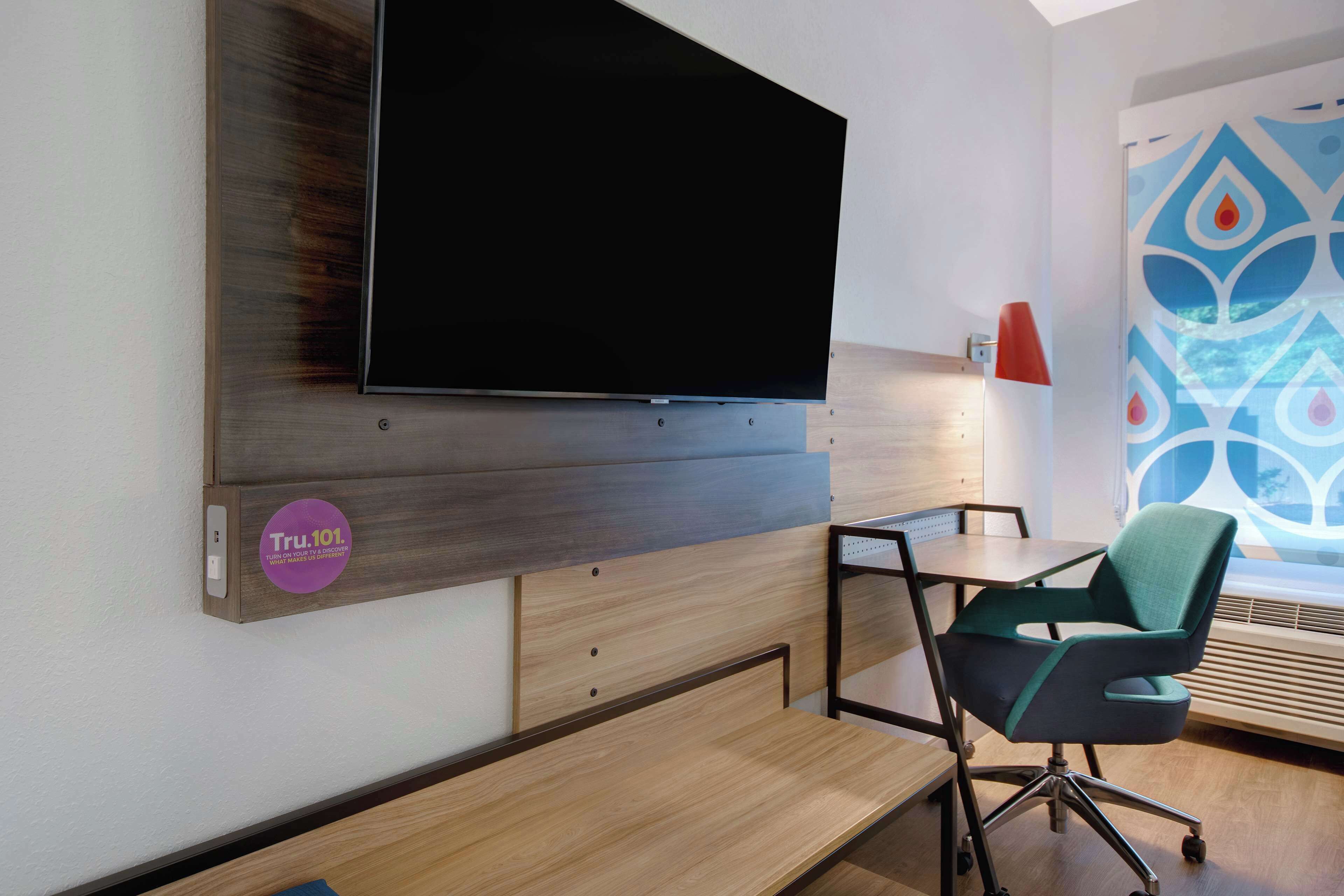 Una habitación moderna con una televisión de pantalla plana montada en un panel de madera, un escritorio, una silla verde y vistas a una ventana con un diseño.