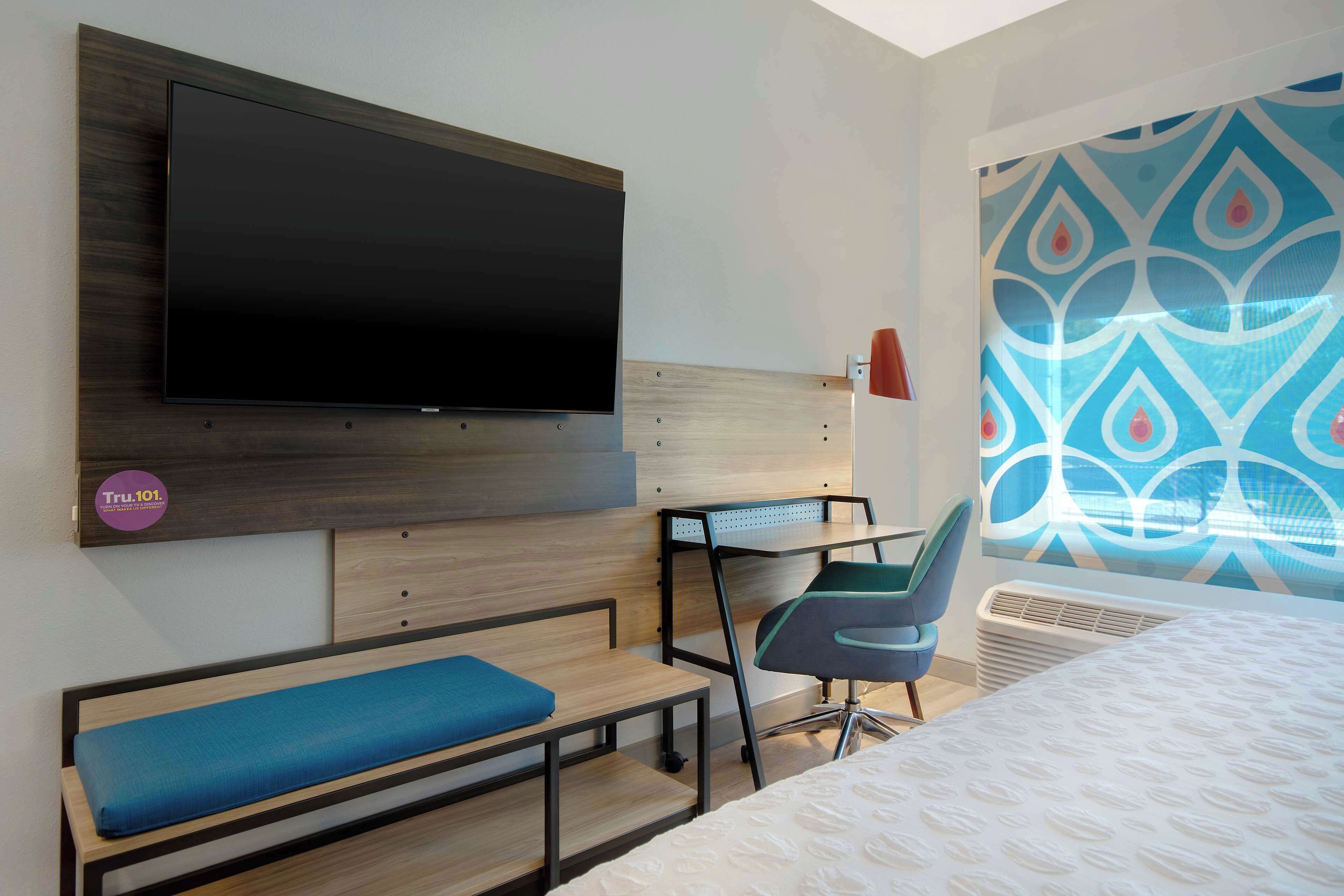 A hotel room with a large flat-screen TV mounted on the wall, a desk with a chair, a bench, and a bed with a patterned bedspread.