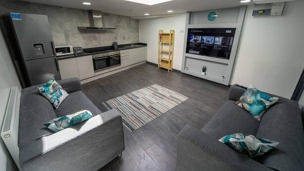 A modern kitchen with a built-in oven, microwave, and a large TV screen. There are two grey sofas with teal cushions and a small wooden ladder. - Homely Studio Next to City Centre (Birmingham)