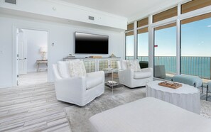 Uma sala de estar moderna com uma televisão de tela grande, sofás brancos, uma mesa de centro e vista para o oceano através de grandes janelas.