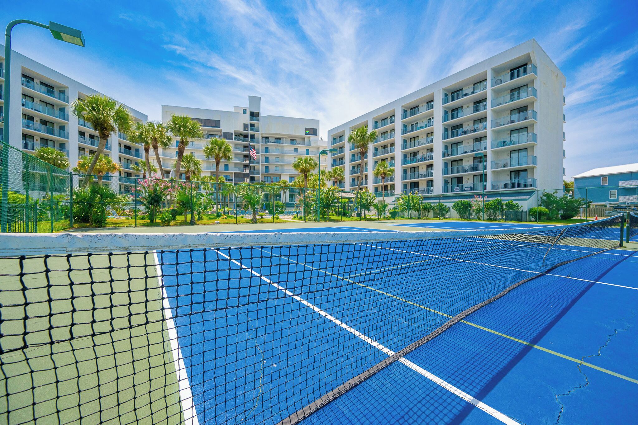 A tennis court with a net in the foreground, surrounded by a fence, and a multi-story hotel building in the background.
