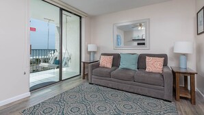 A living room with a grey sofa, patterned rug, and two side tables with lamps. A large window offers a view of a balcony with outdoor furniture.