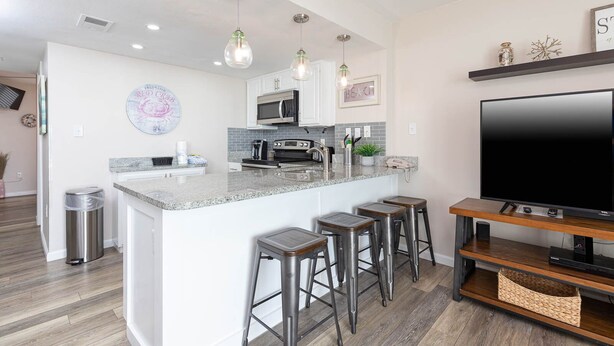 A modern kitchen with a central island, bar stools, a large flat-screen TV, and pendant lighting.