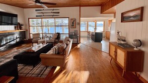 A spacious living area with a wooden ceiling, a sofa set, a dining table, and a kitchen area with a large window.