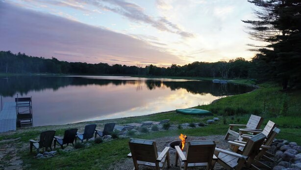 A lakeside seating area with wooden chairs and a fire pit, surrounded by rocks and a view of the lake.