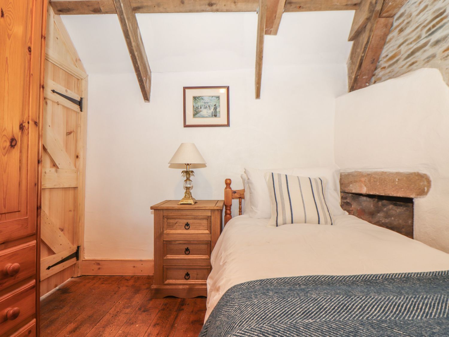 A bedroom with a wooden bed, a nightstand with a lamp, a wooden wardrobe, and a framed picture on the wall.