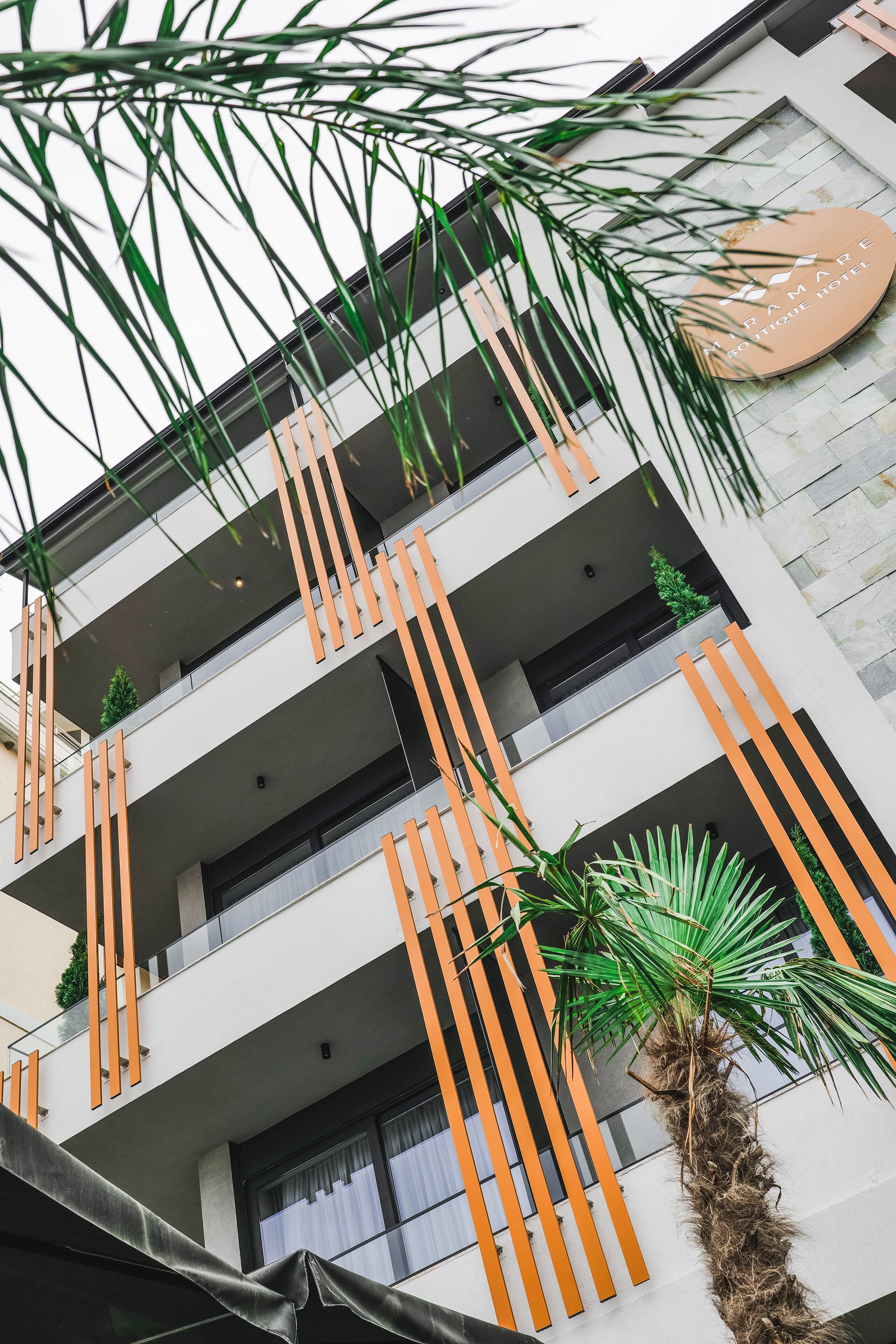 A multi-story building with balconies, orange vertical elements, and a palm tree.
