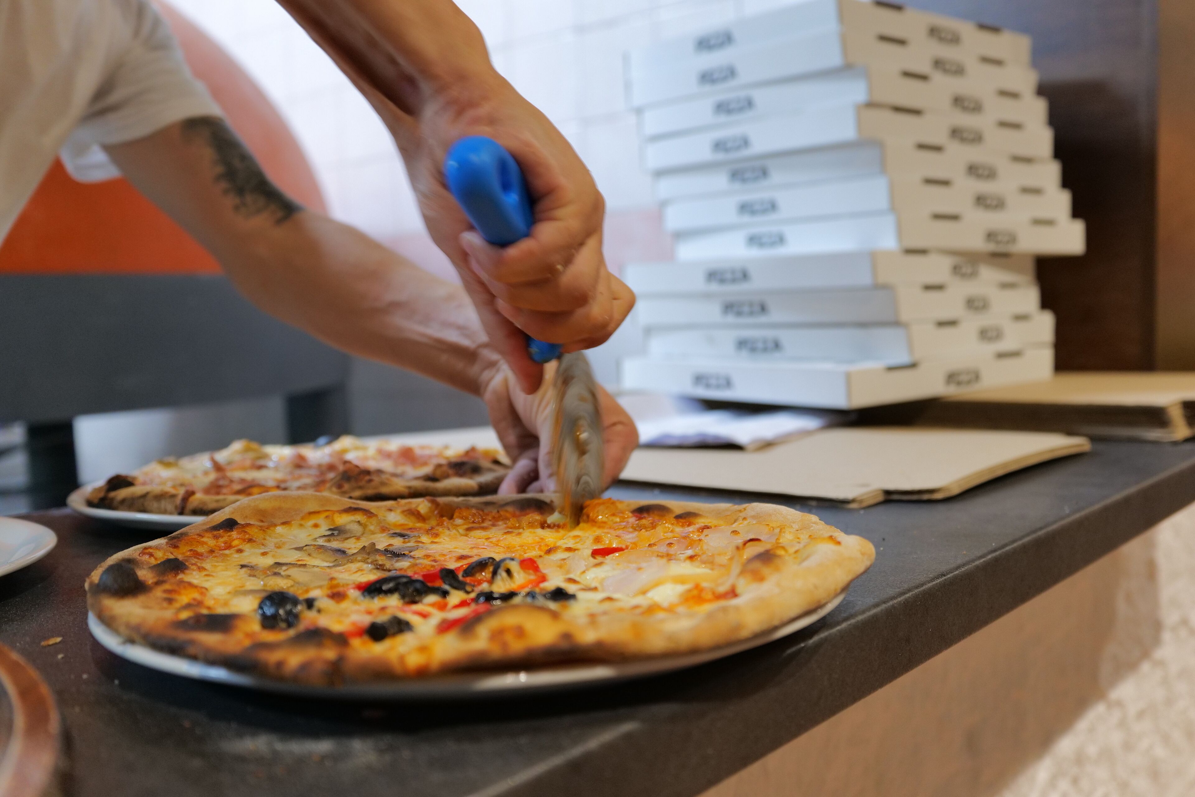 A person cutting a pizza on a counter with a stack of pizza boxes in the background.