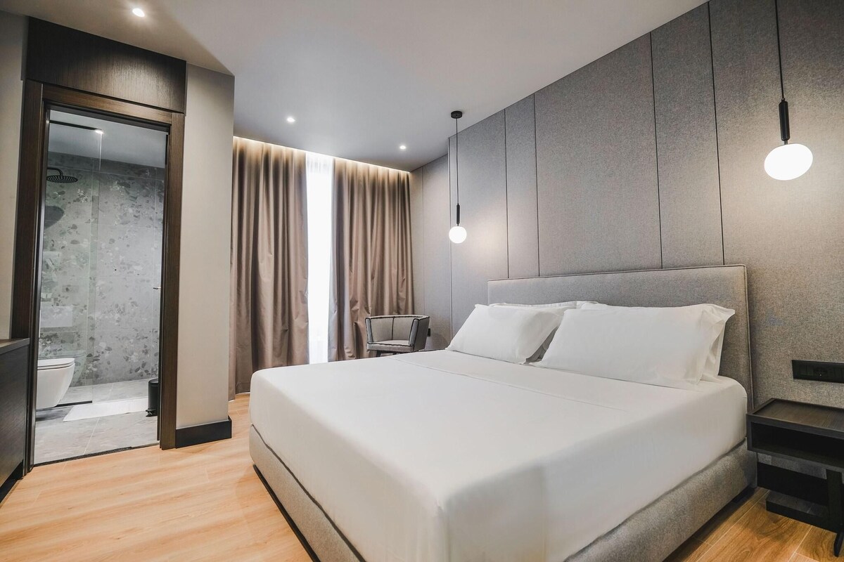 A modern hotel room with a large bed, a bedside table, a chair, a wardrobe, and a bathroom visible through an open door.