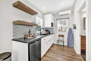 Una cocina compacta con lavavajillas, armarios blancos, un estante de madera y una ventana con persianas.
