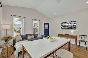 Una sala de estar moderna con un sofá, una mesa de comedor con un cuenco de fruta, un televisor en la pared y una ventana con persianas.