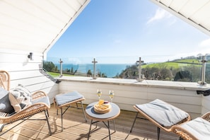 Een balkon met rieten stoelen, een ronde tafel met een kaasplank en glazen, en een prachtig uitzicht op de oceaan.
