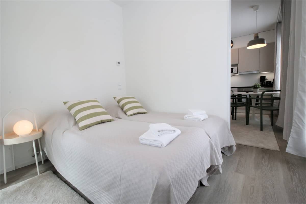 A modern hotel room with two beds, a small desk, and a kitchenette.