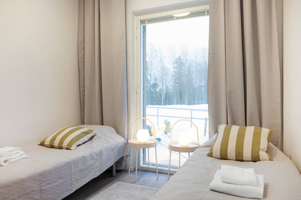 A bedroom with two beds, a window with curtains, and a view of trees outside.